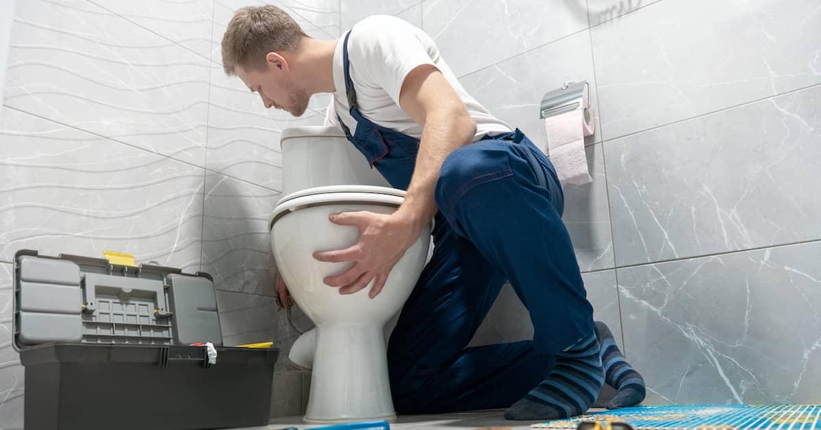 A plumber kneeling on the floor beside a toilet with an open toolbox close by.