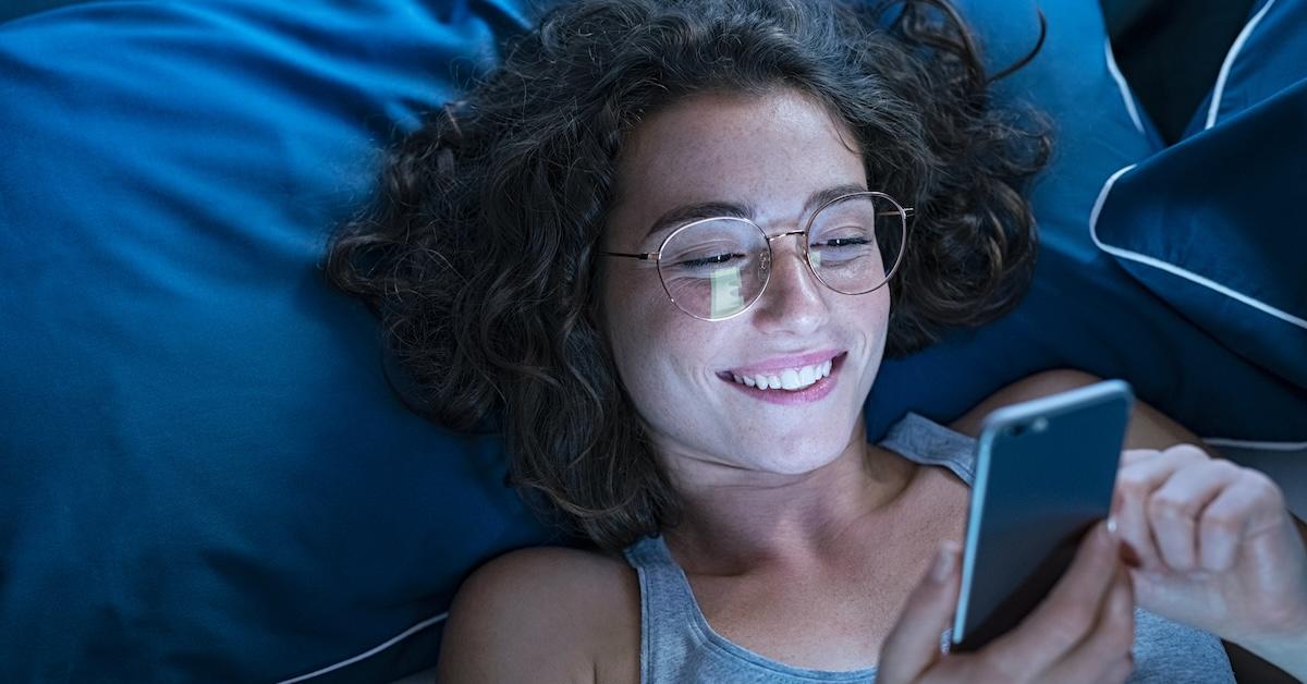 Smiling woman wearing blue light glasses and using smartphone late at night