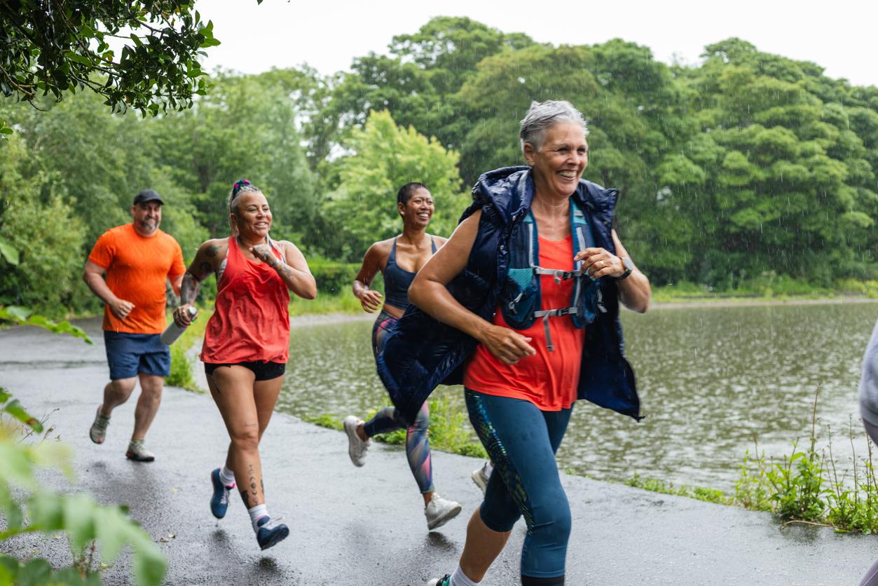 Smiling older adults practice running together in the rain beside trees and a lake.