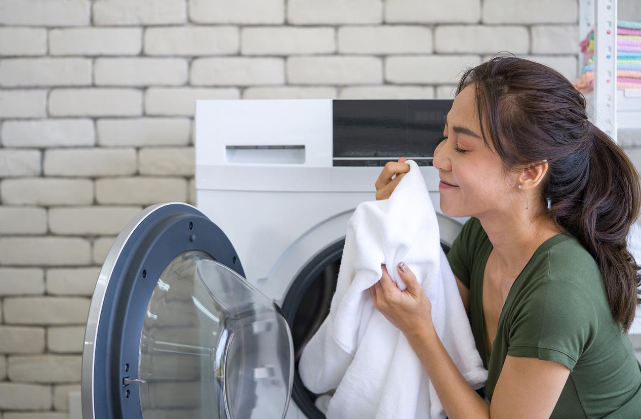 A smiling woman in a green shirt holds a white blanket next to an open dryer.