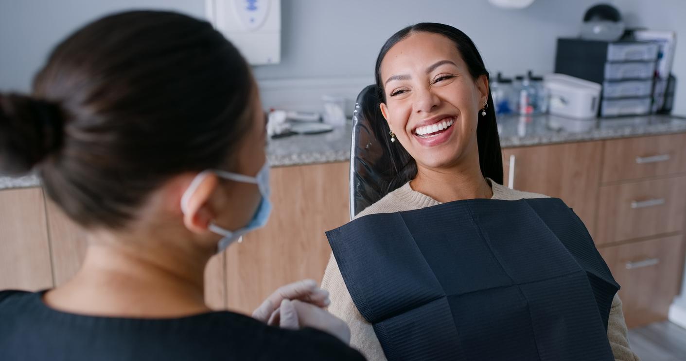 A smiling woman with a black bib lays in a dentist chair while talking to a dental hygienist.