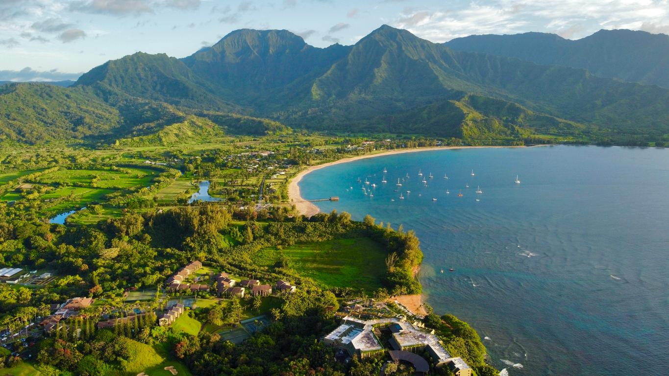 An aerial shot of Hanalei Bay in Kauai featuring mountains, green grass and trees, and a large body of blue water.  