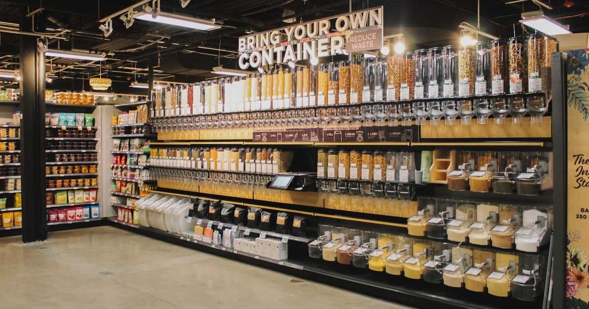 Bulk bins filled with various foods at MOM's Organic Market, with a sign that reads "bring your own container, reduce waste."