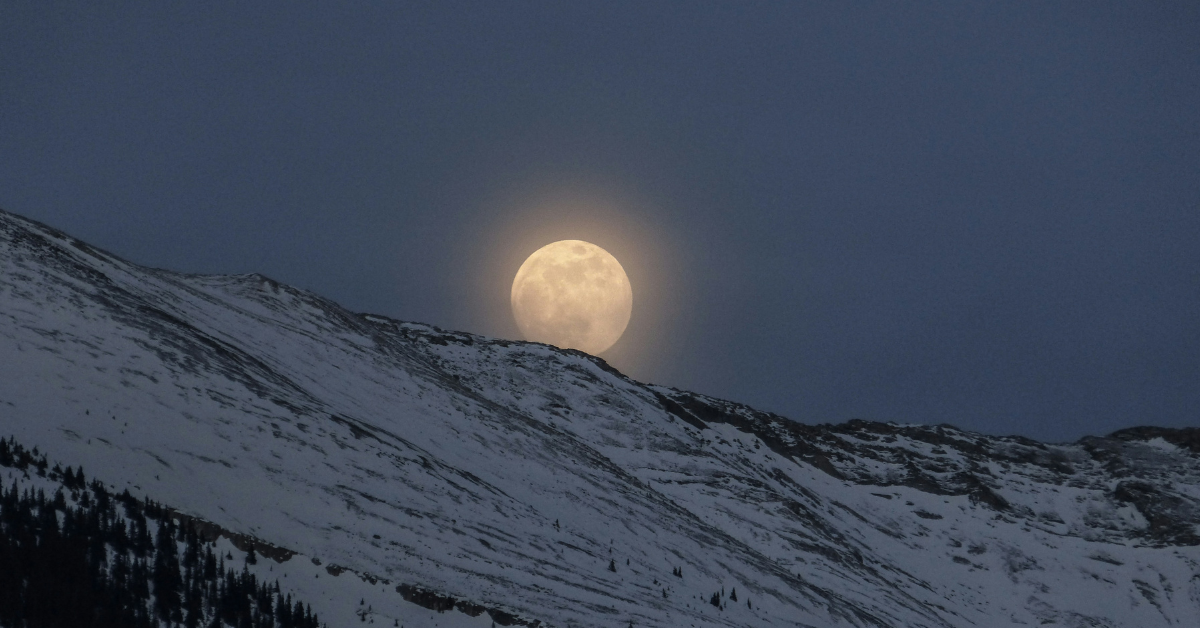 A closeup of a full moon, partially hidden behind a snowy mountainside 