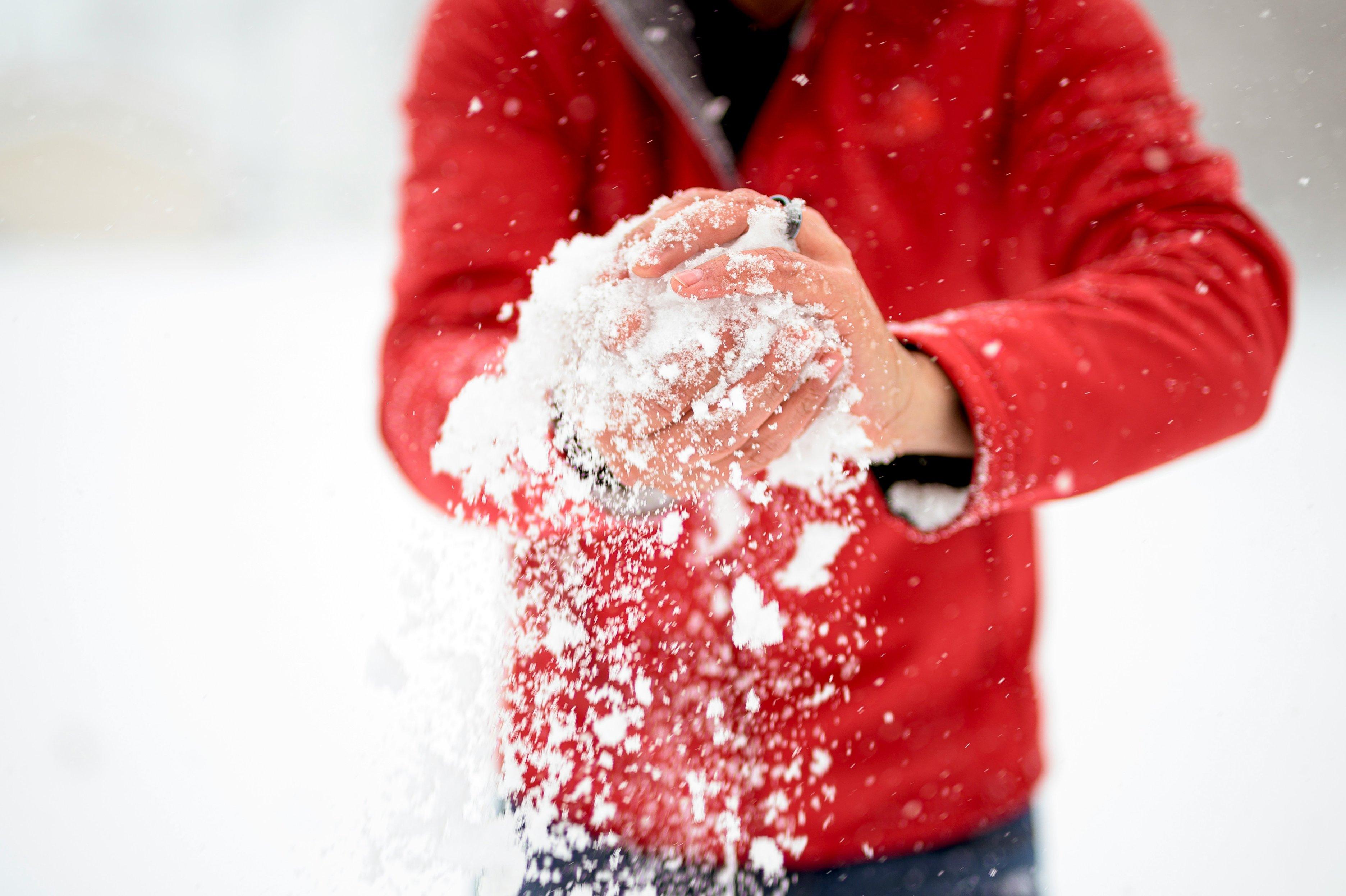 A man in a red winter coat smushes snow together with both hands to make a large snowball.