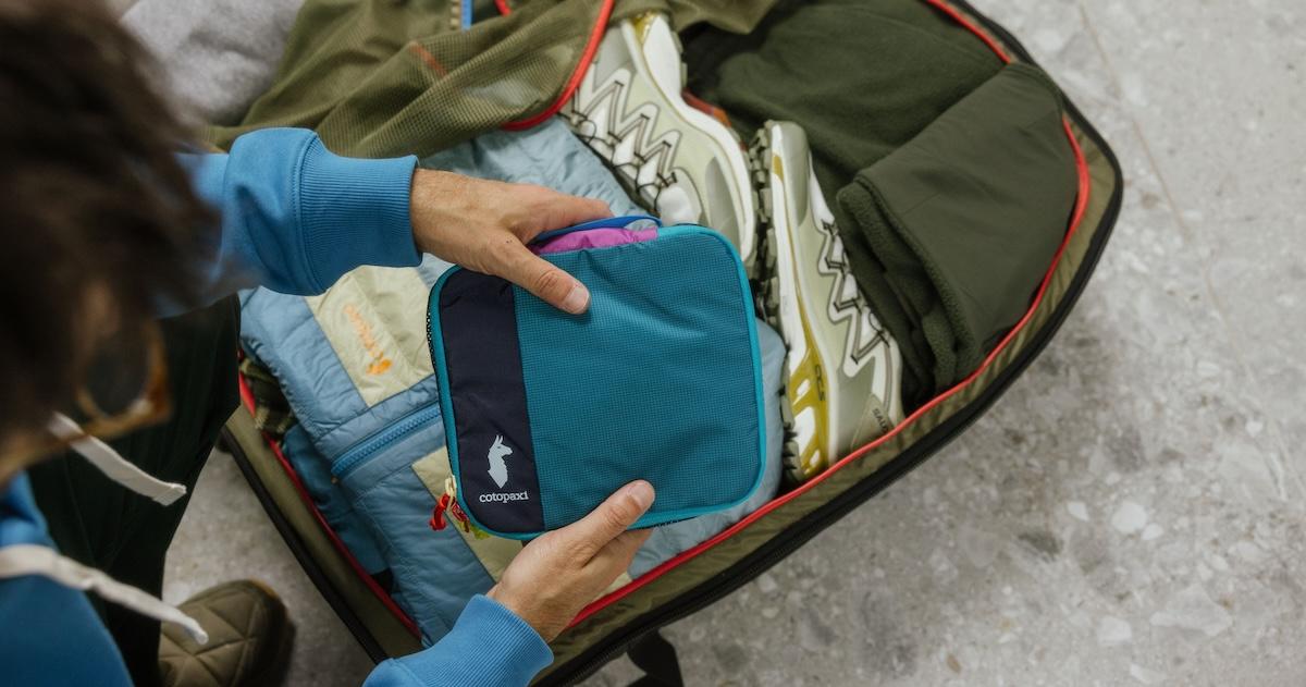 Man holds Cotopaxi packing cube over open suitcase on ground