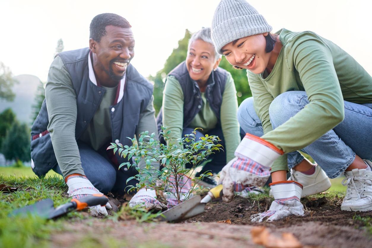 Community members of different ethnic backgrounds smile as they work together to plant a tree in a park.