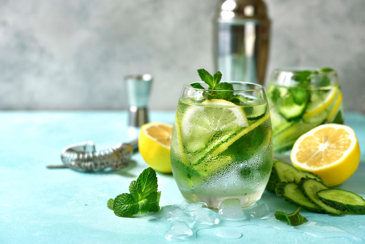 Two glasses of cucumber water garnished with mint and lemon atop a table next to cut cucumbers, lemons, and mint leaves.