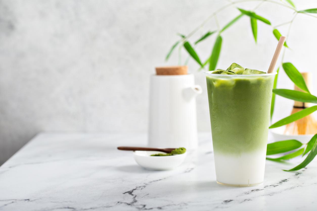 An iced matcha latte on a white table with a plant and matcha powder in the background.