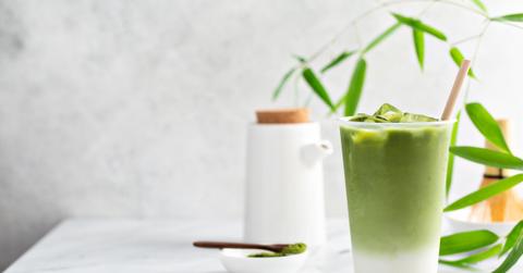 An iced matcha latte on a white table with a plant and matcha powder in the background.