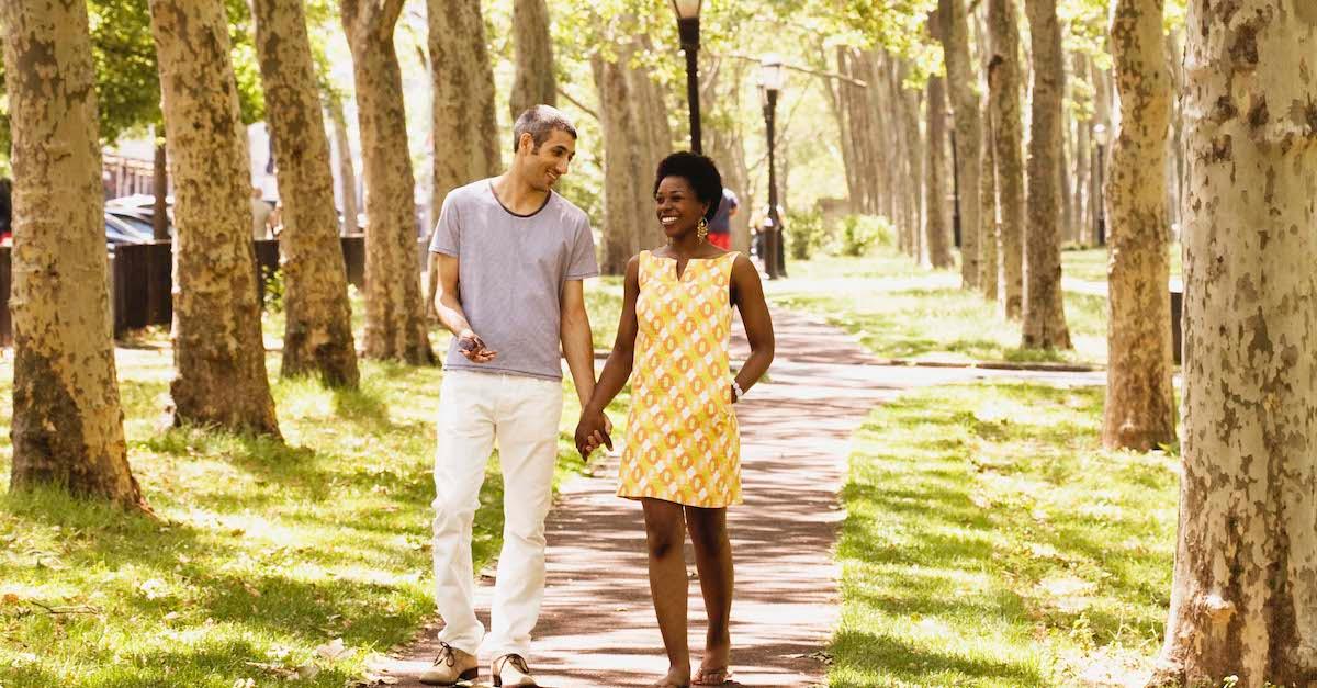 A couple holds hands on a path surrounded by grass, trees, and sunshine.