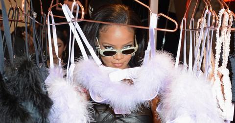 Rihanna wears sunglasses and stands into the camera, while posing with furry bras hanging on a clothing rack