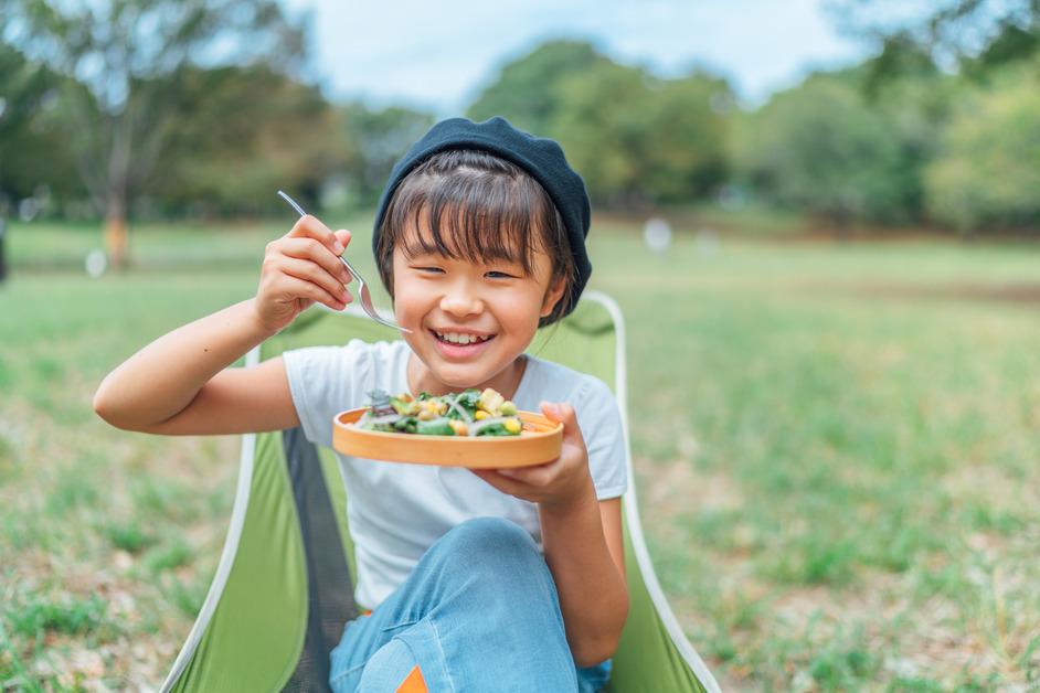 A girl wearing a hat sits outside while eating a plate of vegetables. 