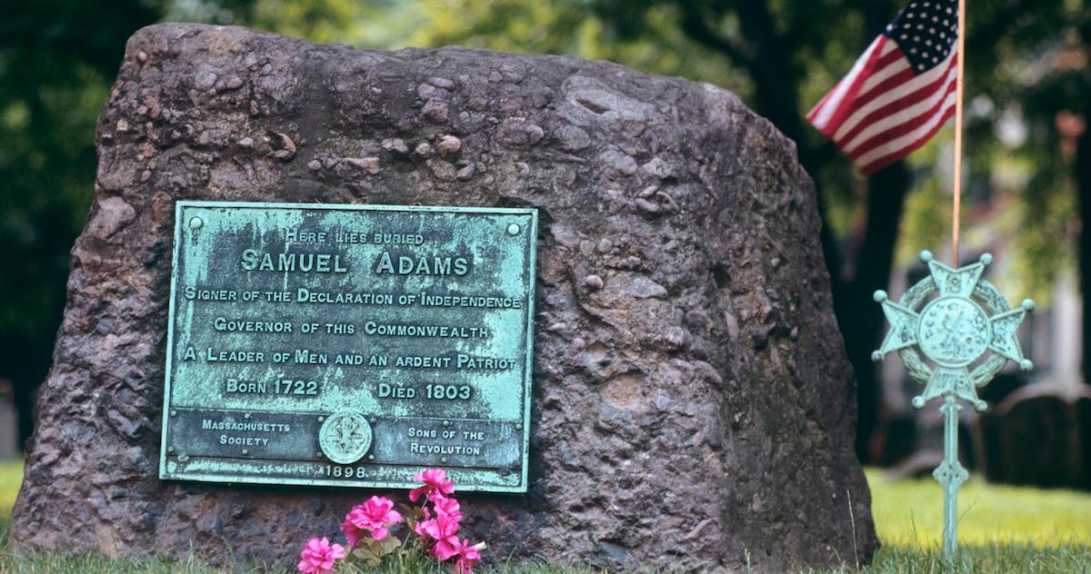 Bronze memorial plaque for Samuel Adams on a rock, next to an American flag. 