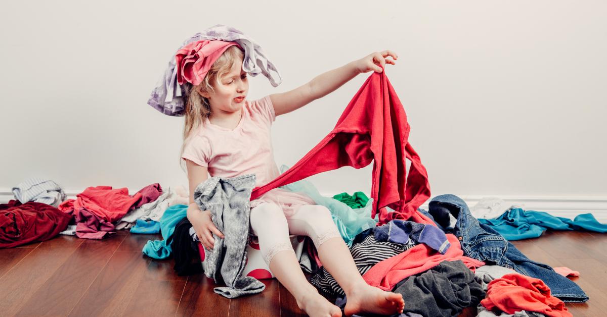 Photo of young blonde girl with items of clothing on her head sits in pile of laundry on hardwood floor