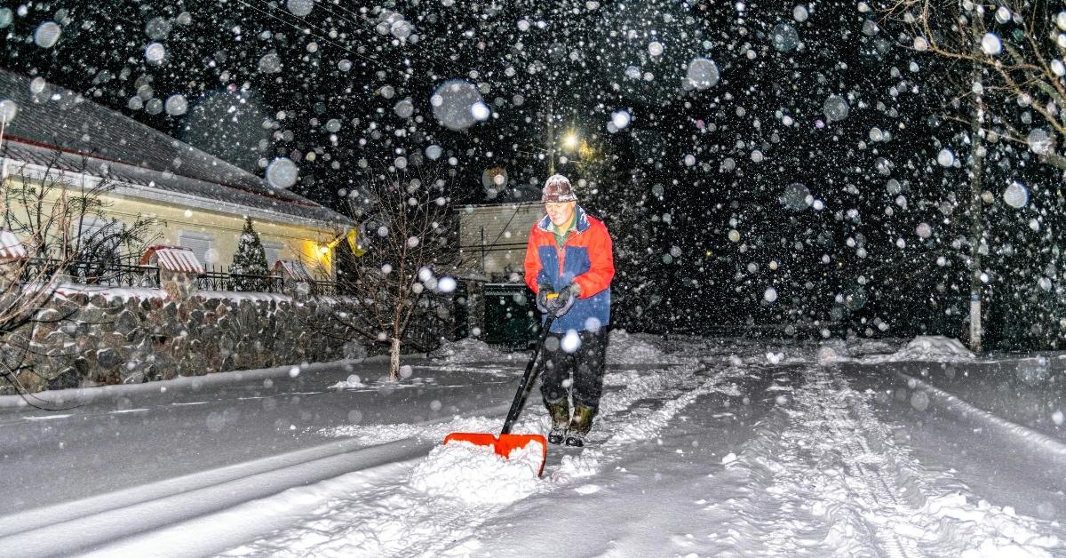 Elderly man in an orange and navy blue jacket shovels snow in his driveway. 