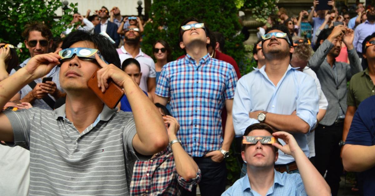 A group of people stand outside while wearing solar eclipse glasses and look up at the sky. 