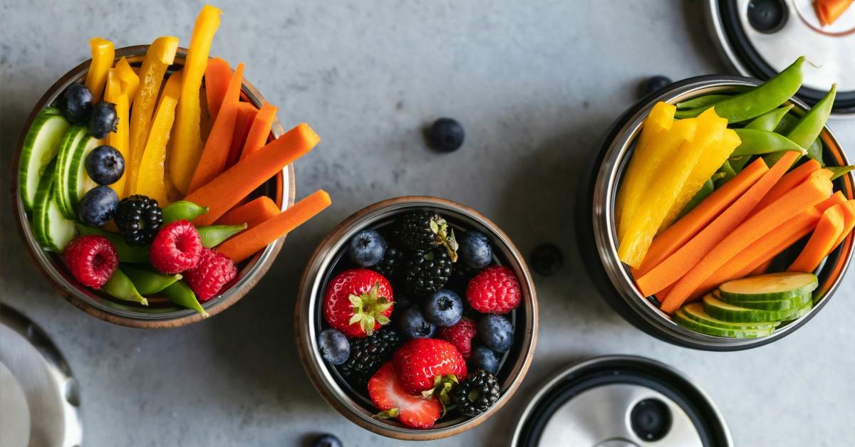 Three metal bowls hold a mix of fruits and vegetables, including carrots, strawberries, and peas