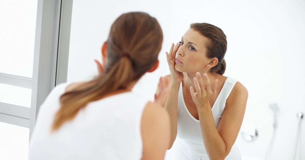 A woman wearing white looks at her face in the mirror.