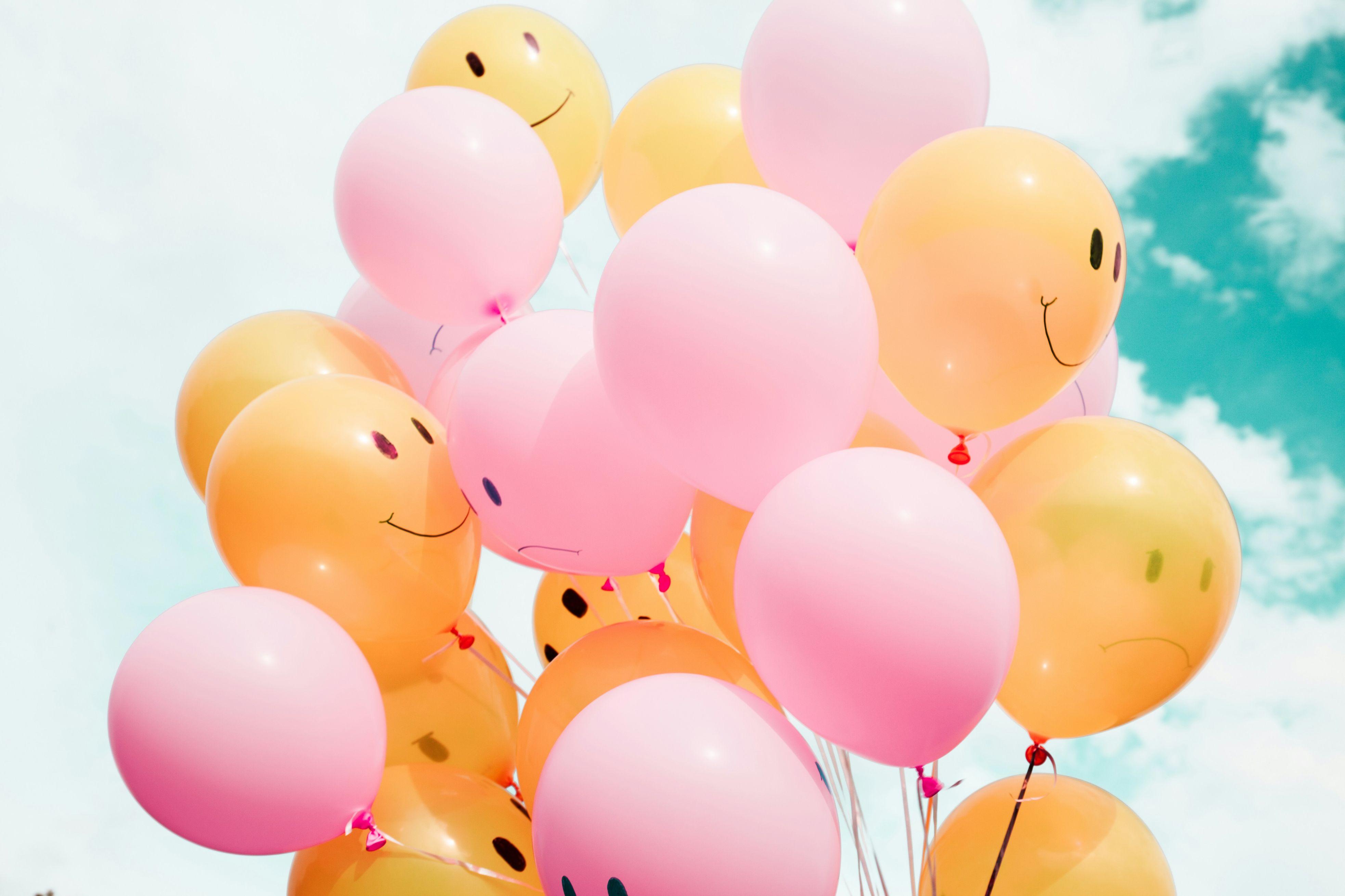 A collection of yellow and pink smiley face balloons appear against a blue cloudy sky.