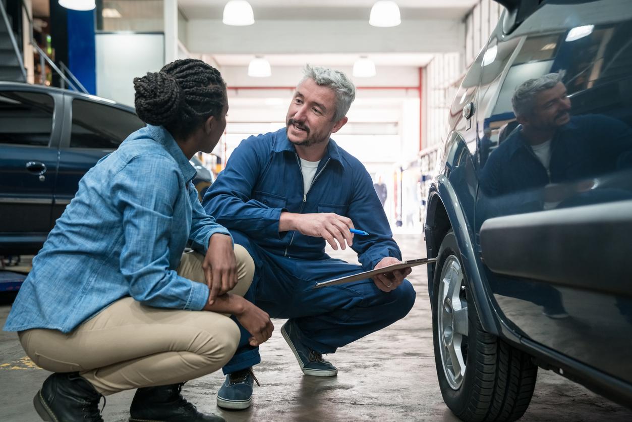 A smiling car mechanic squats beside a customer inside a garage.