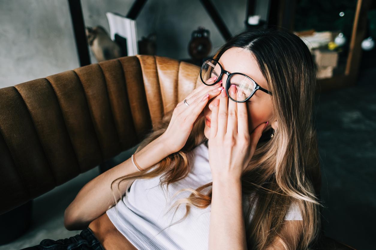 A woman rubs her eyes in pain while laying on a couch.