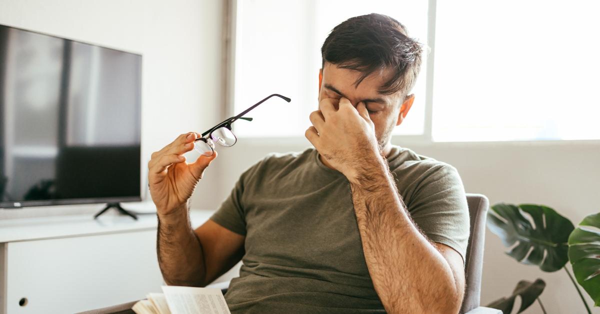 A man in a green shirt pinches the bridge of his nose while holding his glasses to express that he is tired. 