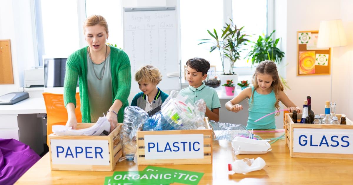 A teacher stands of front of wooden crates labeled "Paper," "Plastic," and "Glass" to teach her students how to sort recycling. 