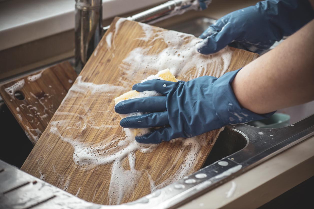 An individual with gloved hands cleans a wooden cutting board with a sponge in a kitchen sink.