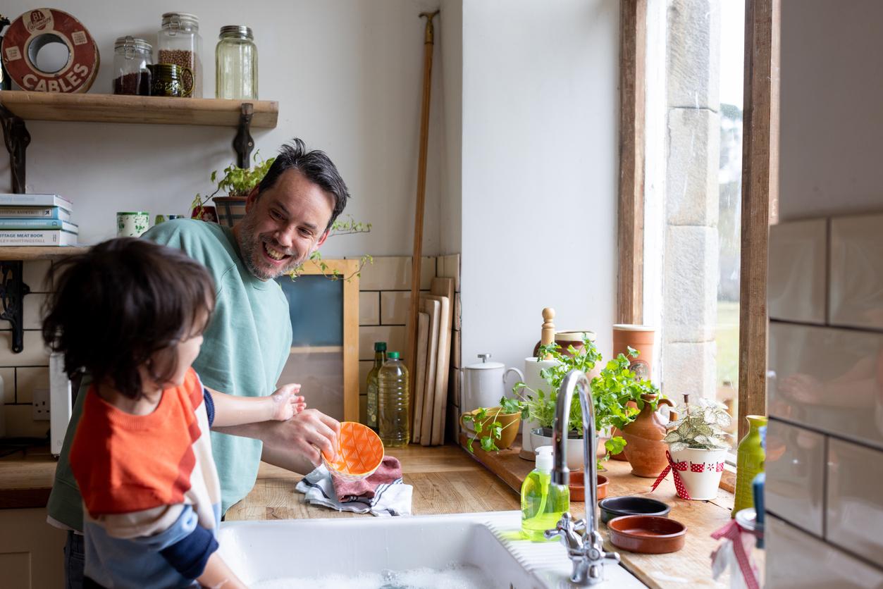 A smiling dad washes dishes with his child on the sink counter of their kitchen.