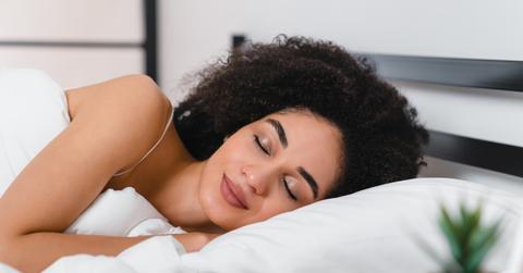 A smiling woman lays with her eyes closed on a white pillow with white sheets and blankets in her bed.