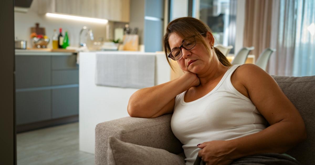 A woman wearing a white tank top sits on the couch resting her head on her hand looking tired. 