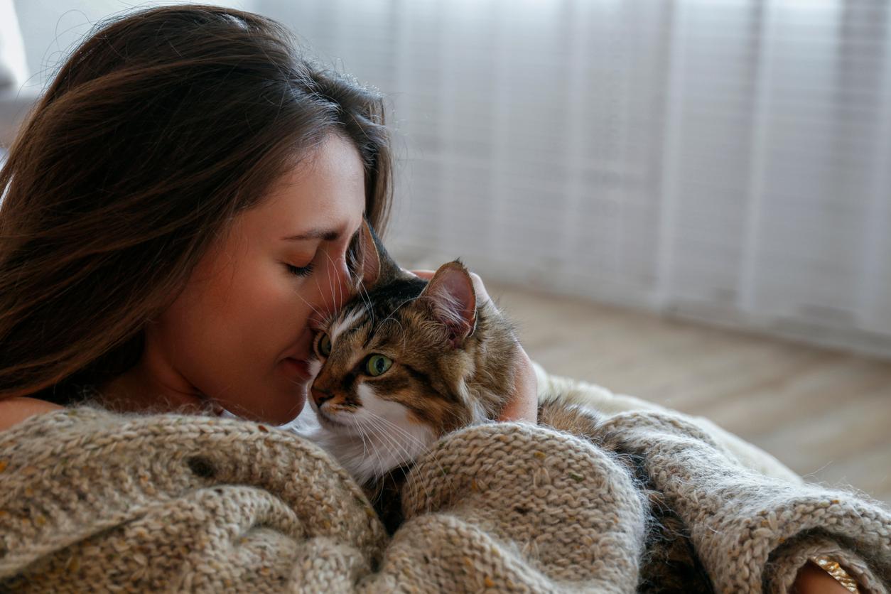 A woman wearing a sweater holds a cat in her arms while bringing her lips and nose to the side of the cat's head.