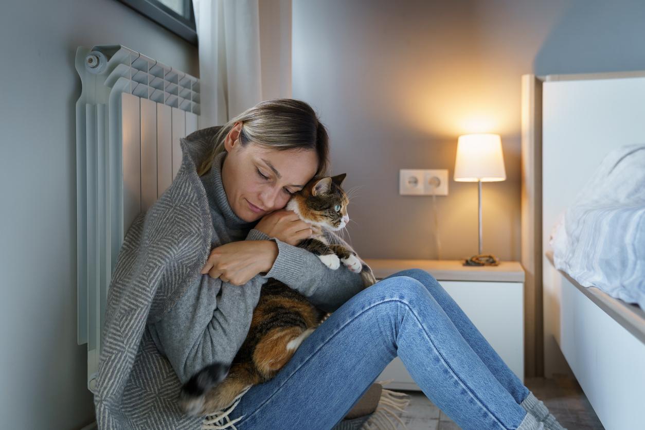 A woman wearing a sweater with a blanket over her shoulders holds a cat close to her chest in a bedroom.