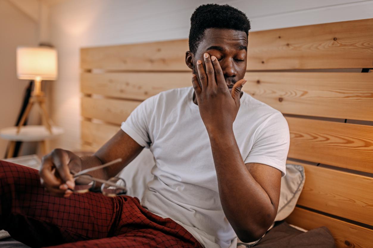 A young man rubs his right eye in pain with his hand while sitting upright in bed.