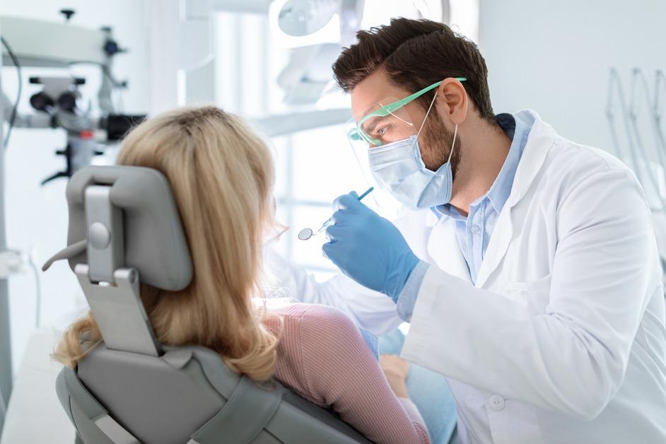 A female patient at the dentist's office sits in a chair as the male dentist wears glasses and a mask while holding tools. 