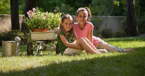 Two young girls sit in a partially shaded yard with flowers ready to be planted.