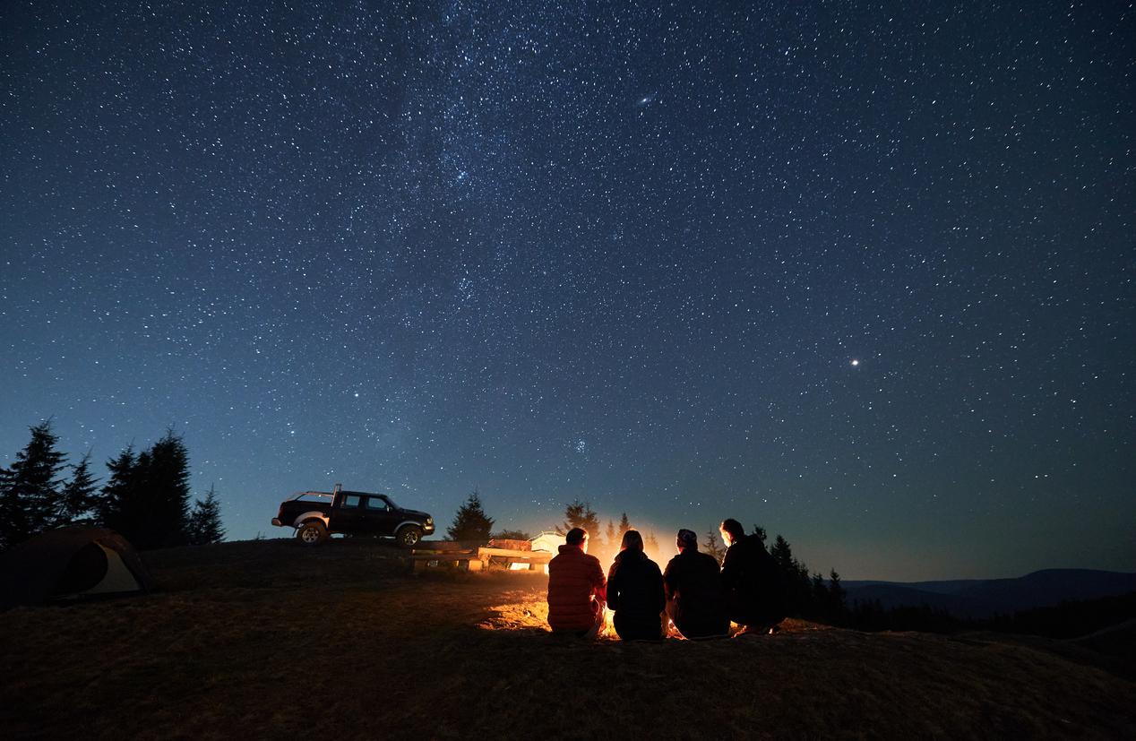 A group of four campers hanging out near a camp fire under a starry night sky.