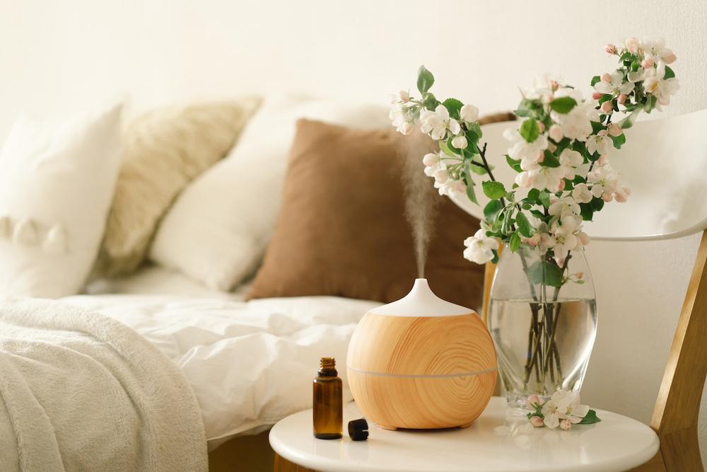 A diffuser sitting on an end table next to flowers and essential oils in a bedroom. 