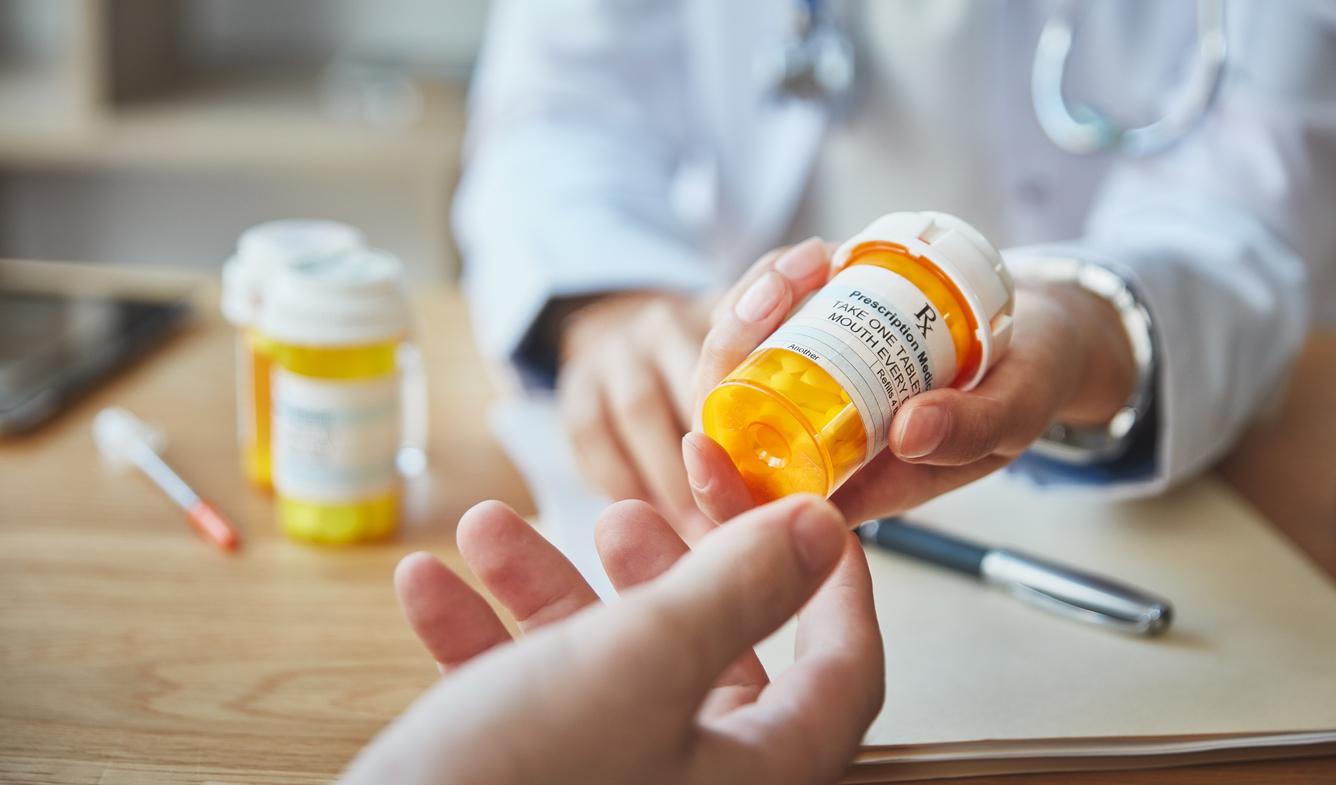 A pharmacist in a white coat hands a prescription pill bottle to a customer.