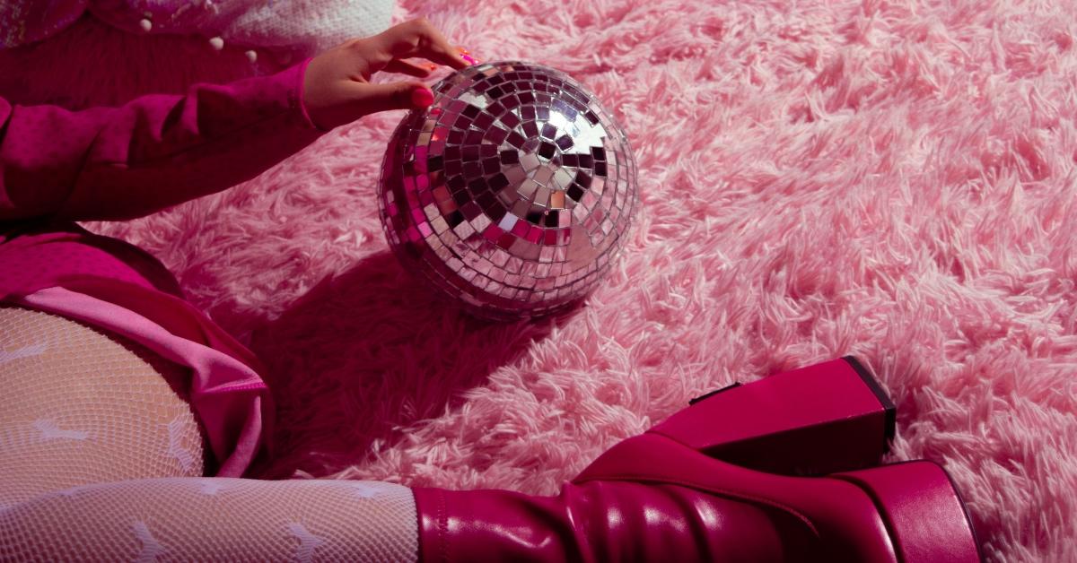 Person wearing a pink outfit and chunky pink heels lays on a pink carpet, holding a mirrorball.