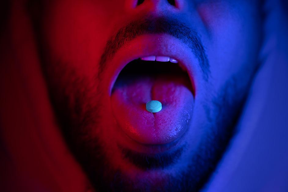 A close-up of a man sticking out his tongue holding a blue pill on it in red and blue lighting. 