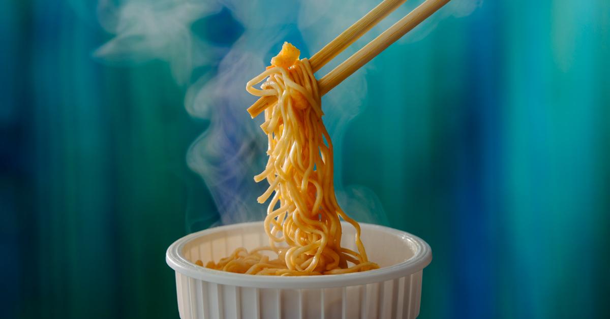 Photo of chopsticks picking up ramen noodles from a cup in front of a blue-green background