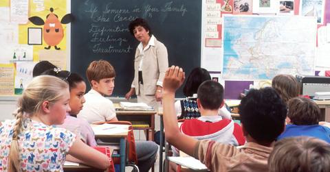 A teacher stands in front of a black board while a student raises their hand