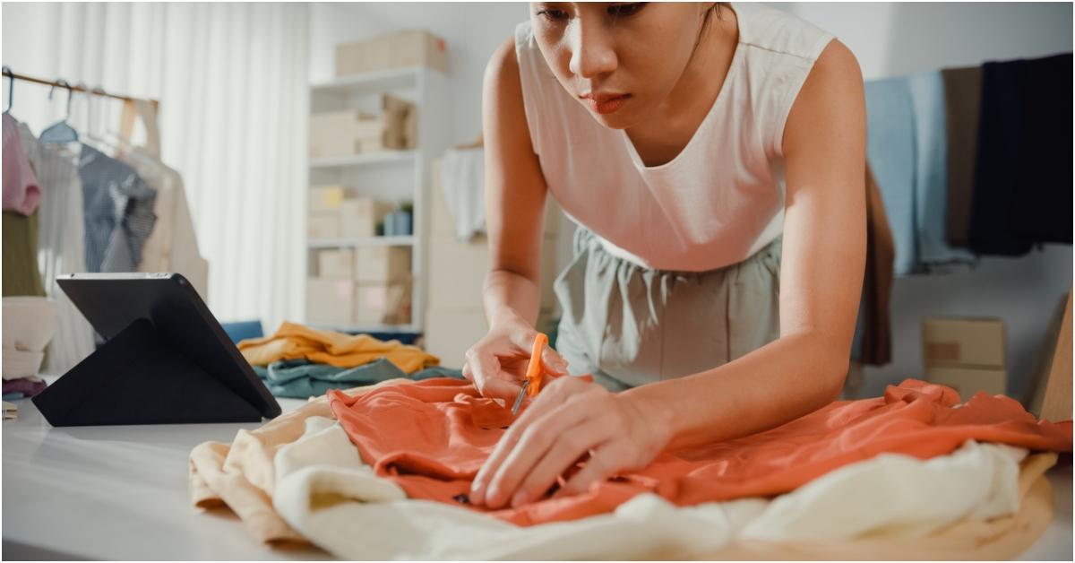 A woman bends over and cuts a shirt with scissors over a pile of clothes next to a tablet on a table. 