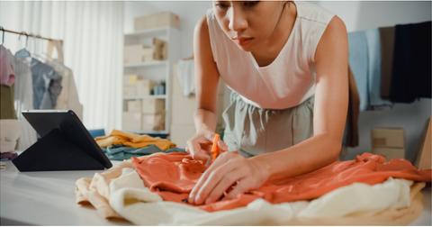 A woman bends over and cuts a shirt with scissors over a pile of clothes next to a tablet on a table.