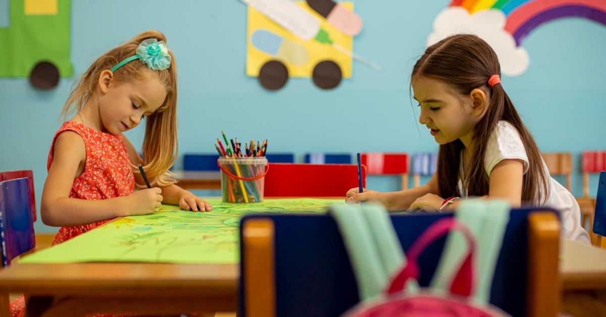 Two young girls sit at a classroom table while using colored pencils on a green sheet of paper. 