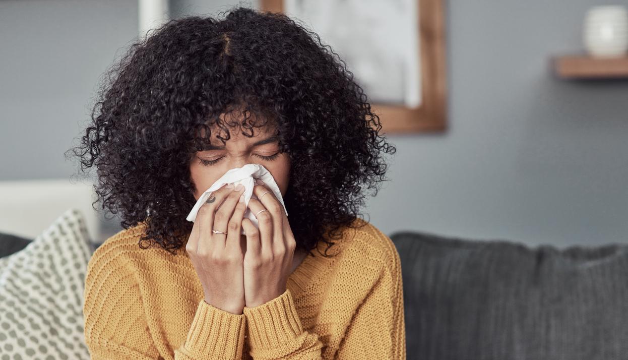 A woman sitting on a couch blowing her nose into a tissue.