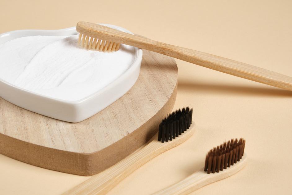 A wooden toothbrush sits in a heart-shaped container of baking soda and two other toothbrushes sit below. 