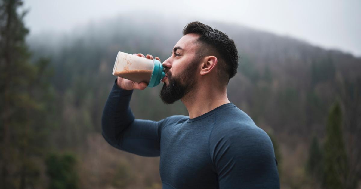 A man wearing a long sleeve shirt stands in front of a mountain while drinking a protein shake.